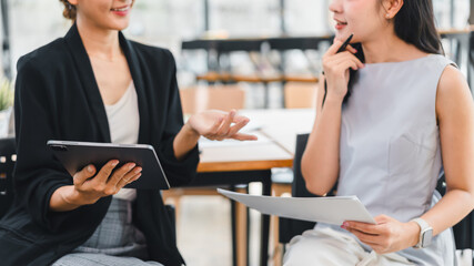 Two businesswomen having friendly discussion while holding documents and tablet in modern office setting, showing engagement and collaboration