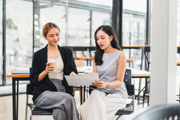 Two young women discussing documents in modern office setting, one holding coffee cup, both dressed...