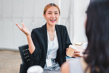 Young woman smiling and holding paper coffee cup while talking to colleague in office setting,...