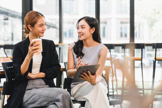 Two young women having friendly conversation in bright modern cafe, one holding coffee cup and other using tablet, sharing ideas with smiles and relaxed posture