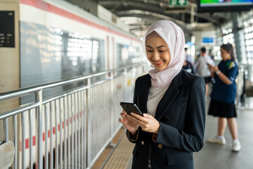 Muslim asian woman using smartphone while waiting on platform at train station in the city....