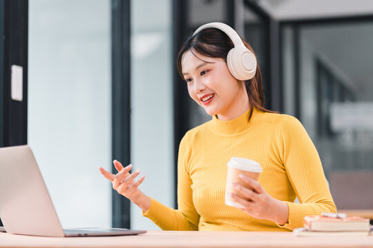 Young woman wearing headphones and yellow sweater is smiling while holding coffee cup and engaging in video call on her laptop in modern office setting