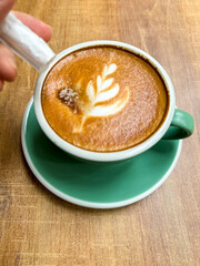 A hand pours sugar into a hot latte with beautiful leaf latte art in a green ceramic cup. Captured on a wooden table with warm tones, emphasizing cozy café vibes and sweet coffee moments.

