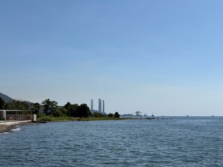 Obraz premium Serene ocean view contrasting with industrial elements; a power plant and mountains visible across the water, alongside a natural coastline. 