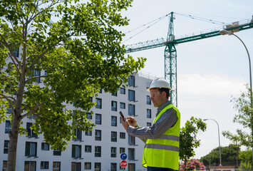 Professional male construction worker with safety helmet and reflective vest and safety glasses using tablet at a construction site with building progress in the background