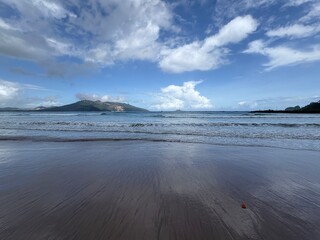 Wet sandy beach with ocean waves and distant green mountains under a blue sky