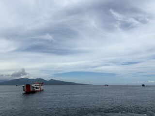 Boats anchored in calm water near a distant port or coastline under a cloudy sky.