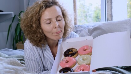 Happy woman with closed eyes licking lips while lying on bed and looking at box of fresh colorful...