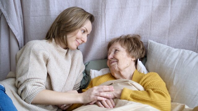 Young woman holding hands of her elderly mother lying in bed, providing comfort and support during illness or difficult time, concept of family, love, and caregiving
