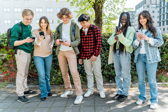 Group of diverse high school students using smartphones outdoors during break in urban school setting