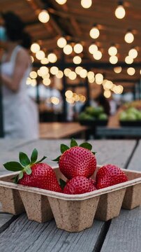 Vertical screen, ripe strawberries lie in an eco friendly cardboard container on a wooden table at a blurred farmers market with warm string lights and vendors in the background