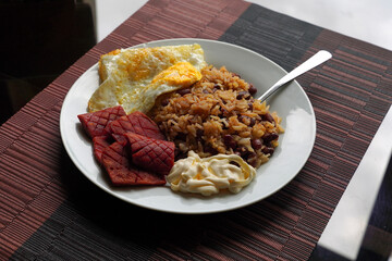 Gallo pinto, a typical Costa Rican dish with rice, red beans, sausage, cream, and a fried egg, and a glass of coffee on a table with a nature background.