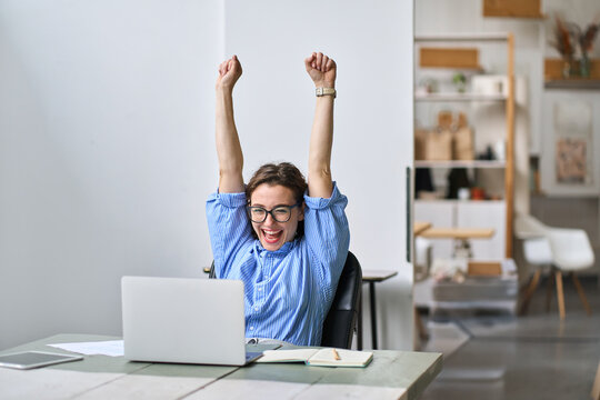 Excited office worker winner using laptop computer celebrating goal achievement. Happy young woman student or employee winning online getting good news in email raising hands feeling euphoric.