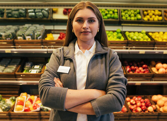 Portrait of a young woman grocery store worker standing with hands crossed against shelves with fruits and vegetables