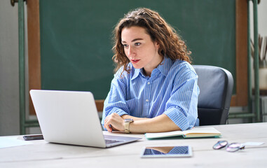 Female online teacher looking at laptop talking on virtual distance class in classroom or webinar web training working in office. Young business woman leading hybrid conference online video call.
