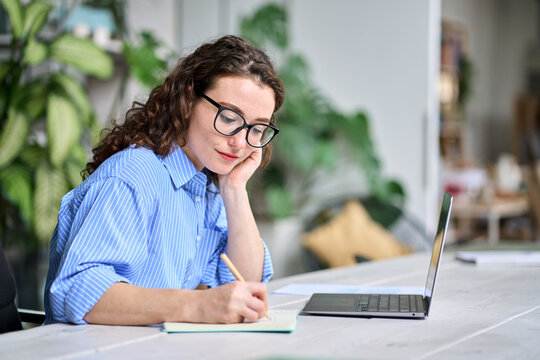 Female employee watching online webinar training, doing research, managing work tasks sitting at desk in office. Busy young business woman or student using laptop working or learning taking notes.
