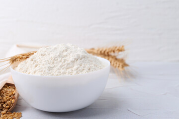 Wheat flour in bowl, grains and spikes on white table, closeup. Space for text
