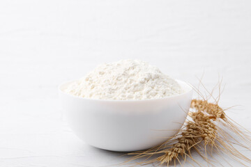Wheat flour in bowl and spikes on white table, closeup