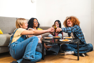 Four cheerful young multiethnic friends are toasting with beer bottles while sitting on the floor around a small table and eating pizza at home