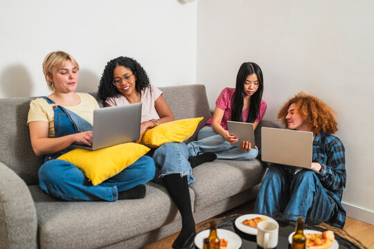 Four young multiethnic students using laptops and digital tablets while sitting on a sofa and on the floor in a modern apartment