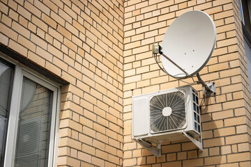 Satellite dish and air conditioning unit mounted on exterior wall of brick building during sunny afternoon