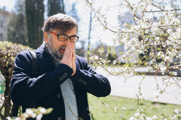 Senior man suffering from allergy symptoms near blooming tree
