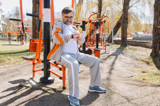Senior man checking smartwatch sitting on outdoor gym equipment - Powered by Adobe