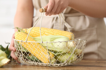 Woman holding basket with fresh corn cobs on wooden table