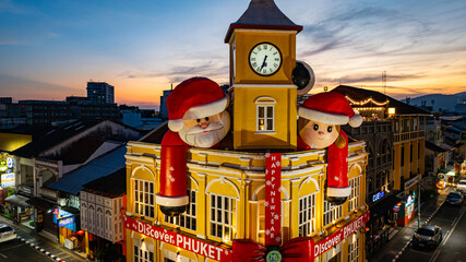 Phuket,Thailand-December,30,2024: A vibrant scene featuring a historic yellow clock tower adorned with giant Santa Claus and Mrs. Claus decorations during a dramatic evening sky. Happy New Year 2025
