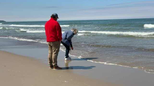 A man and a woman walk along the Baltic Sea beach, carefully searching for pieces of amber washed up on the shore. They bend down occasionally to pick up small golden fragments among the sand and seaw
