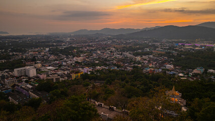 A scenic view of a town nestled among lush green mountains at sunset, with a temple structure perched on the hillside and a vibrant orange sky creating a warm, tranquil atmosphere.