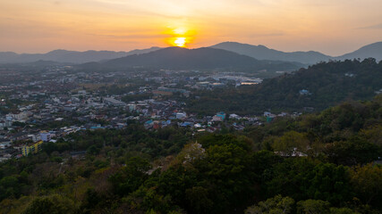 A scenic view of a town nestled among lush green mountains at sunset, with a temple structure perched on the hillside and a vibrant orange sky creating a warm, tranquil atmosphere.