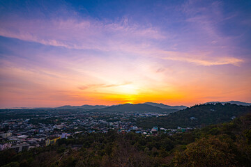 Fototapeta premium A scenic view of a town nestled among lush green mountains at sunset, with a temple structure perched on the hillside and a vibrant orange sky creating a warm, tranquil atmosphere.