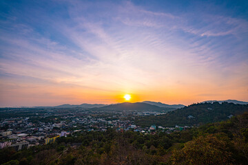 A scenic view of a town nestled among lush green mountains at sunset, with a temple structure perched on the hillside and a vibrant orange sky creating a warm, tranquil atmosphere.