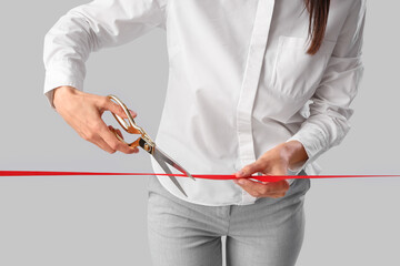 Young businesswoman cutting red ribbon on grey background