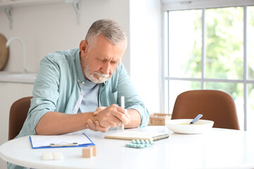 Senior man suffering from Parkinson syndrome writing on notebook at table in kitchen