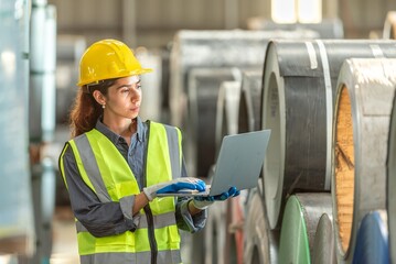 Female engineer wea safety helmet and vest inspecting large industrial metal coils with a laptop in...