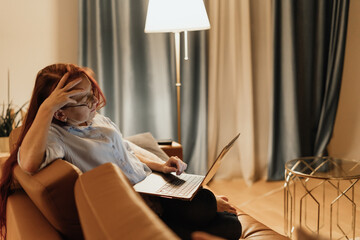 Exhausted woman with laptop touches forehead while sitting on sofa symbolizing cognitive overload screen anxiety and mental fatigue caused by information excess constant online connectivity at home
