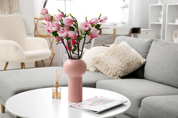 Vase with beautiful magnolia flowers and magazines on table near grey sofa in modern living room