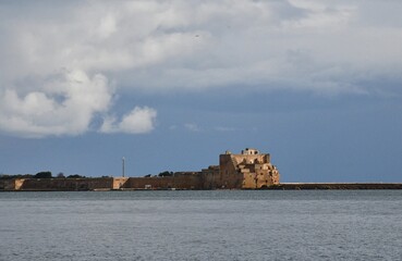 Sunlight falls on ruined stone fortifications on a harbour breakwater. The sky is overcast and the water is grey. The fort and defensive wall are of sandstone.