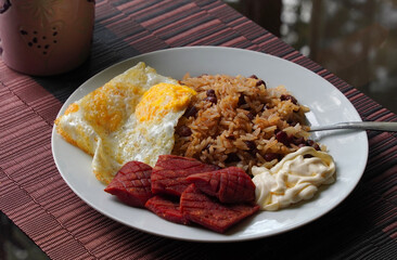 Gallo pinto, a typical Costa Rican dish with rice, red beans, sausage, cream, and a fried egg, and a glass of coffee on a table with a nature background.