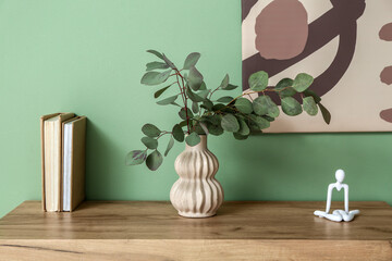 Vase with eucalyptus branches, statuette and books on table near green wall in room, closeup