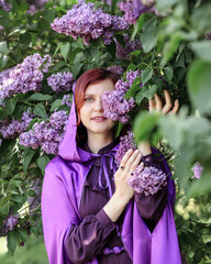 A beautiful young woman with red hair, against a background of blooming lilacs, in a cloak and...