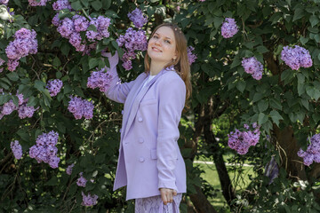 A beautiful young blonde woman, against a background of blooming lilacs, in a lilac trouser suit....