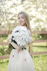 A young beautiful blonde girl in long white dress poses near cherry blossom tree in spring garden,...