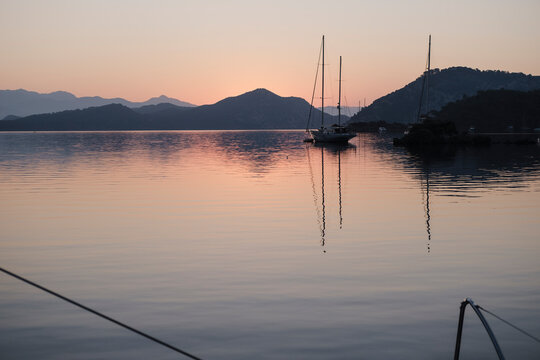 scenic view of morning calm tranquil sunrise from the boat deck at anchor stop, rising sun, mountains, sea with light reflections, twilight colors, sailing boats, serene harbour