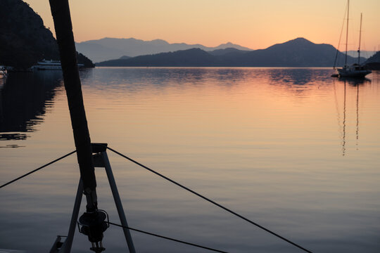scenic view of morning calm tranquil sunrise from the boat deck at anchor stop, rising sun, mountains, sea with light reflections, twilight colors, sailing boats, serene harbour