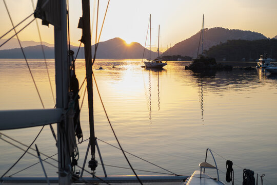 scenic view of morning calm tranquil sunrise from the boat deck at anchor stop, rising sun, mountains, sea with light reflections, twilight colors, sailing boats, serene harbour