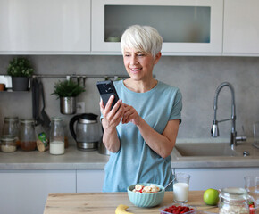 Portrait of a happy senior mature mid aged woman having a healthy breakfast after fitness exercise training and using a smartphone, looking at phone in the morning at home
