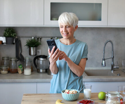 Portrait of a happy senior mature mid aged woman having a healthy breakfast after fitness exercise training and using a smartphone, looking at phone in the morning at home